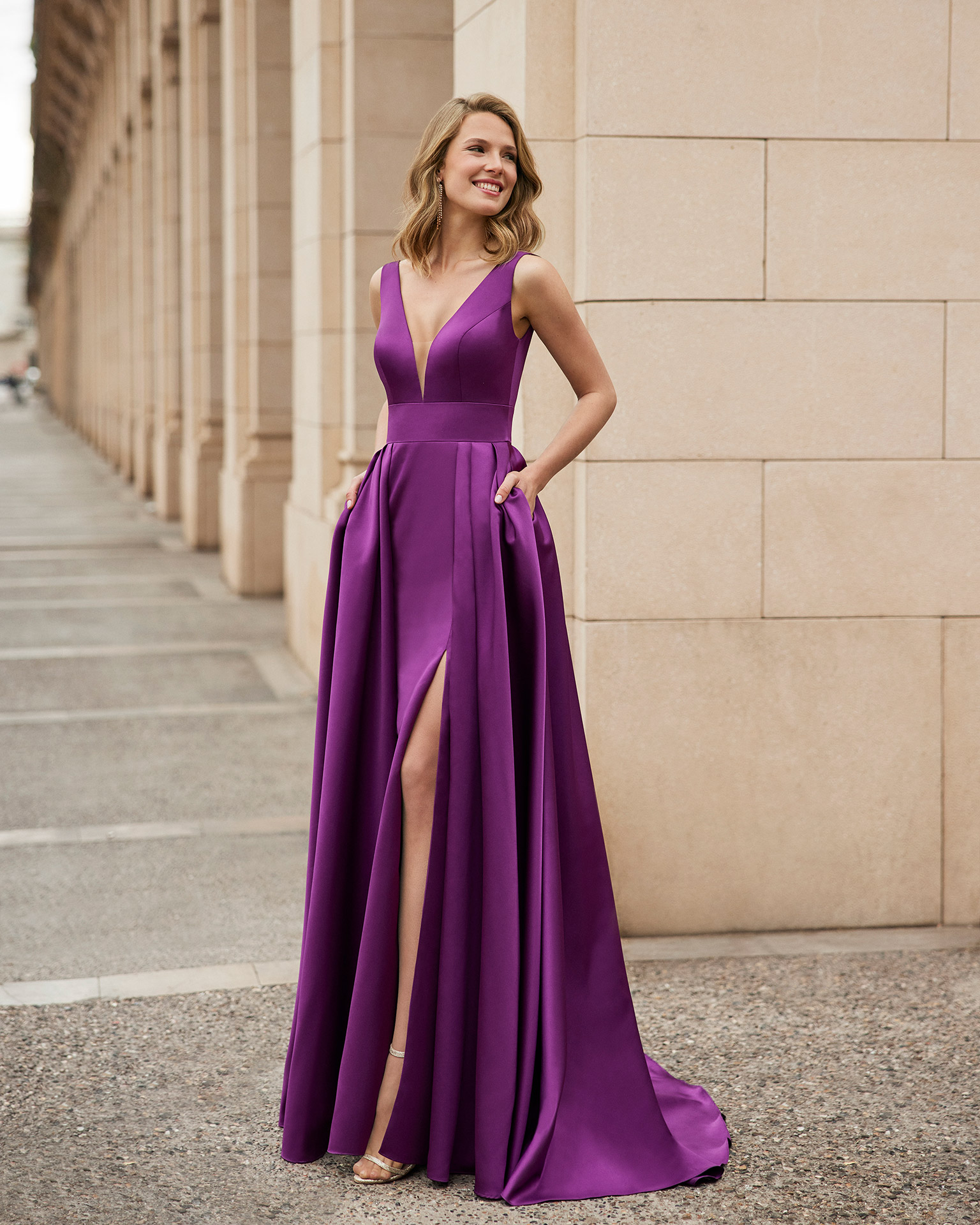 Long evening dress made of satin. Marfil Barcelona design with a V-neckline, and a pleated skirt with a slit. MARFIL_BARCELONA.