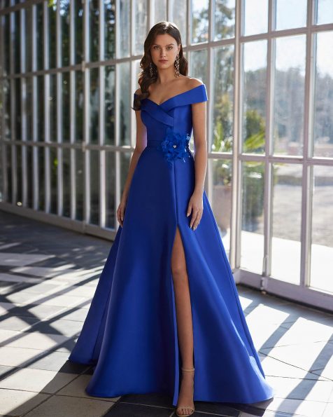 Long evening dress crafted in mikado. Showcase an off-shoulder neckline, sleeveless bodice, skirt with a side slit, and a flower embellishment. A Marfil Barcelona look. MARFIL_BARCELONA.